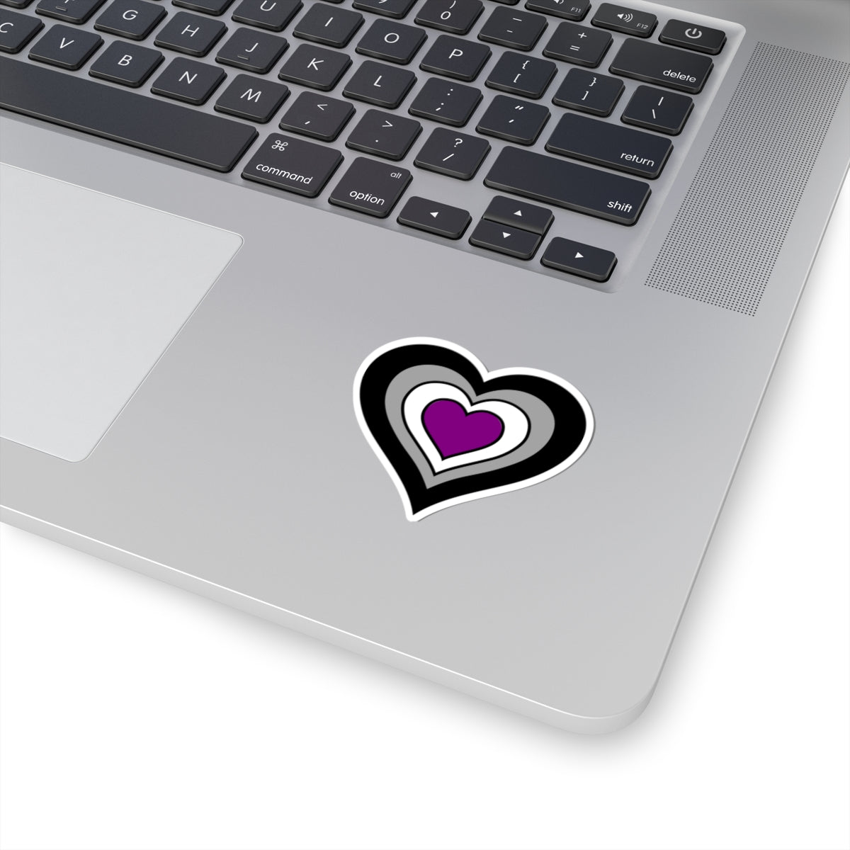 Close-up of the bottom right corner of a silver laptop featuring a 2 inch by 2 inch pride flag heart sticker in an Asexual flag style applied to the surface.