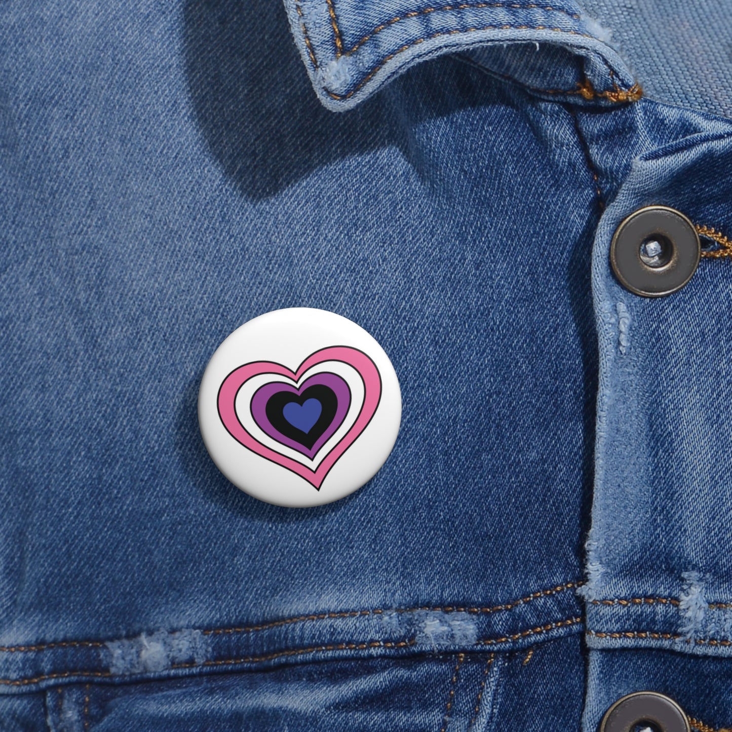 Close-up of a white circular pride button featuring a nested heart design inspired by the Genderfluid pride flag in pink, white, purple, black and blue, attached to a blue denim jacket at the right chest with the button clearly visible.