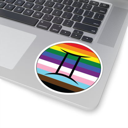 Close-up of the bottom right corner of a silver laptop featuring a 4 inch by 4 inch circular zodiac sticker with a centered Gemini glyph on a progress pride flag style background applied to the surface.