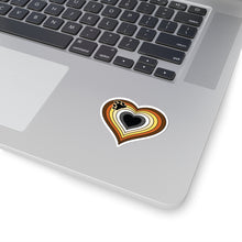 Close-up of the bottom right corner of a silver laptop featuring a 2 inch by 2 inch pride flag heart sticker in a Bear pride flag style applied to the surface.