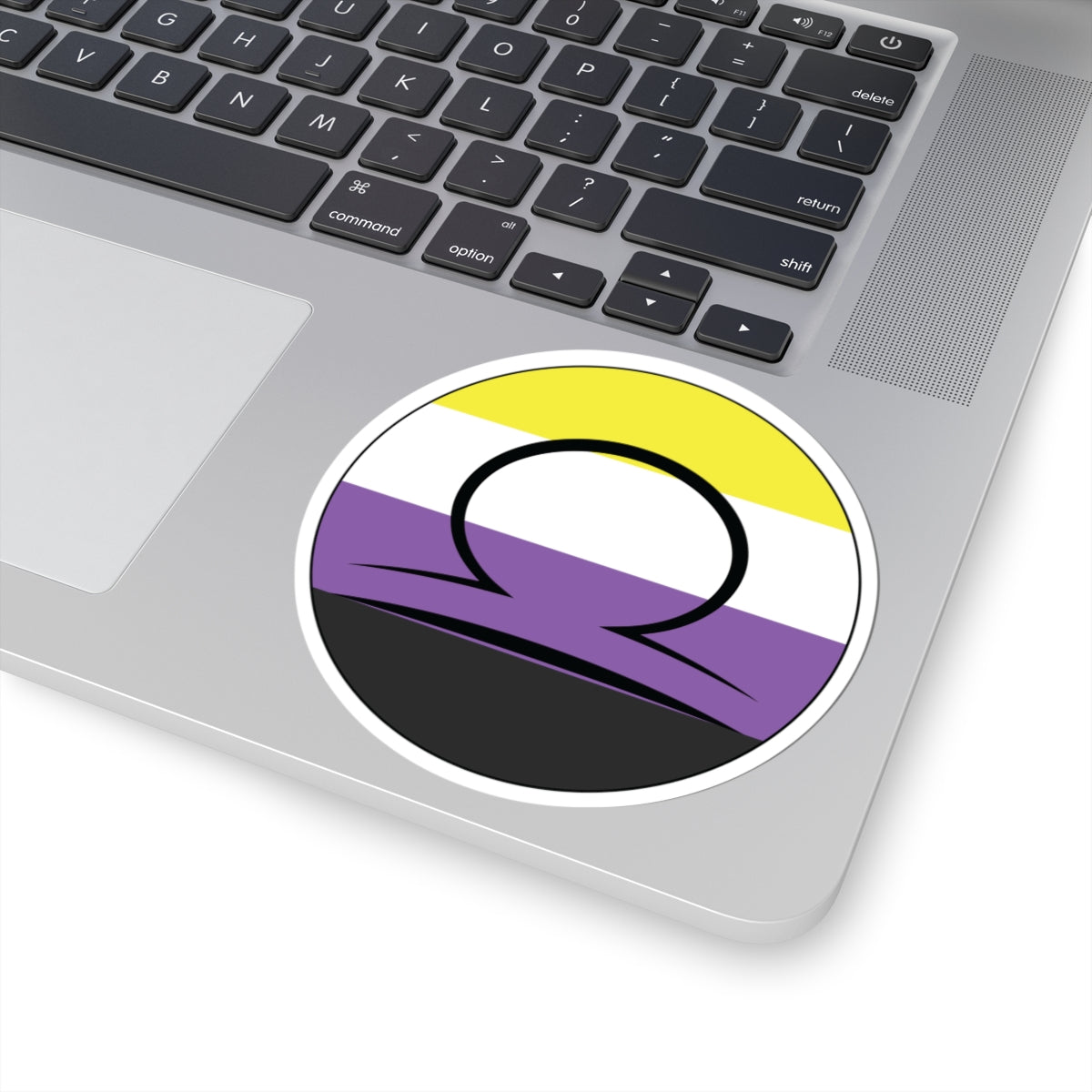 Close-up of the bottom right corner of a silver laptop featuring a 4 inch by 4 inch circular zodiac sticker with a centered Libra glyph on a Nonbinary pride flag style background applied to the surface.