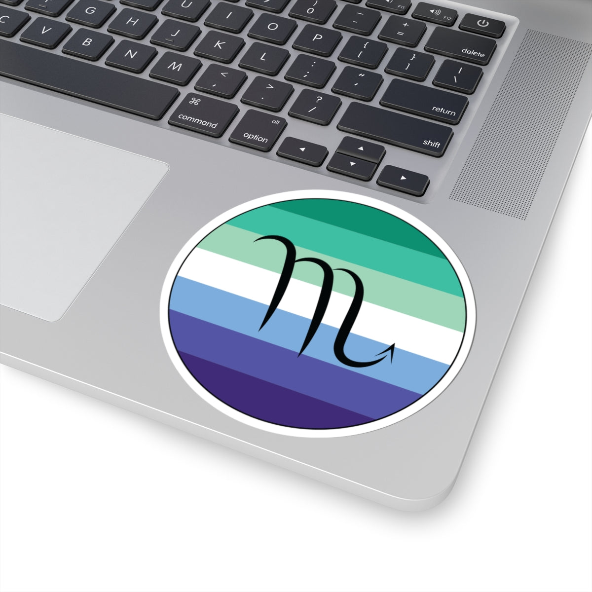 Close-up of the bottom right corner of a silver laptop featuring a 4 inch by 4 inch circular zodiac sticker with a centered Scorpio glyph on a Gay Men pride flag style background applied to the surface.