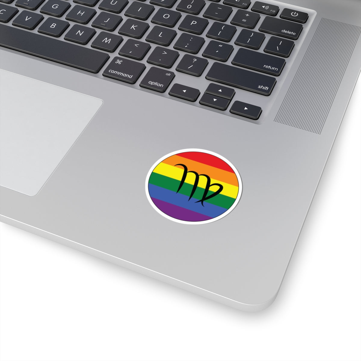 Close-up of the bottom right corner of a silver laptop featuring a 2 inch by 2 inch circular zodiac sticker with a centered Virgo glyph on a Rainbow pride flag style background applied to the surface.