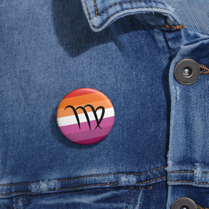 Close-up of a white circular pride button featuring the Virgo zodiac glyph centered over a lesbian pride flag background in oranges, white, and pinks attached to a blue denim jacket at the right chest with the button clearly visible.