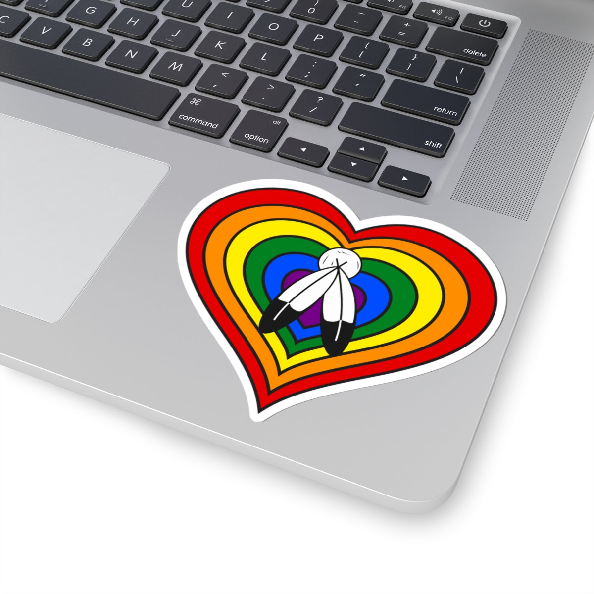 Close-up of the bottom right corner of a silver laptop featuring a 4 inch by 4 inch pride flag heart sticker in a Two-Spirit flag style applied to the surface.