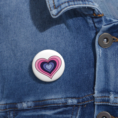 Close-up of a white circular pride button featuring a nested heart design inspired by the Omnisexual pride flag in pinks and purples, attached to a blue denim jacket at the right chest with the button clearly visible.