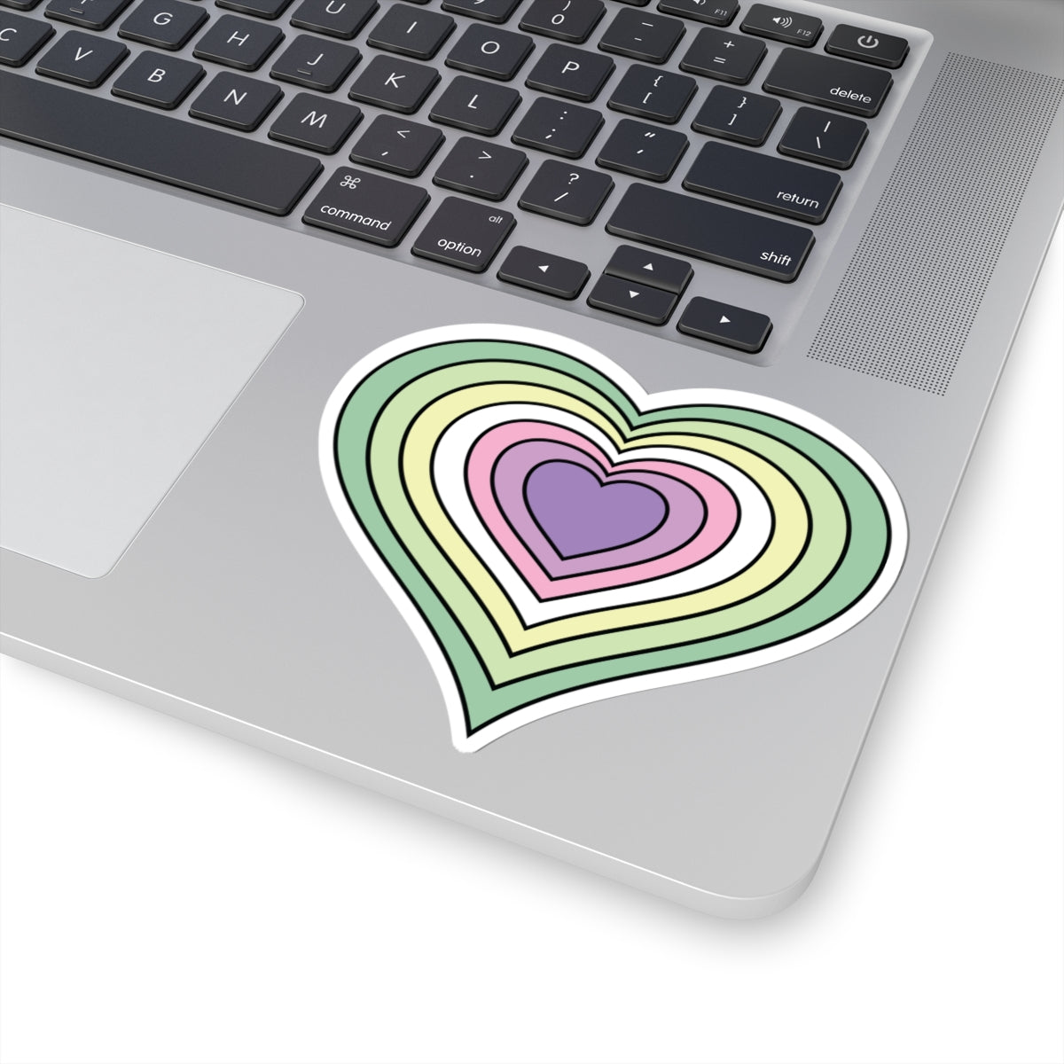 Close-up of the bottom right corner of a silver laptop featuring a 4 inch by 4 inch pride flag heart sticker in a Genderfae flag style applied to the surface.