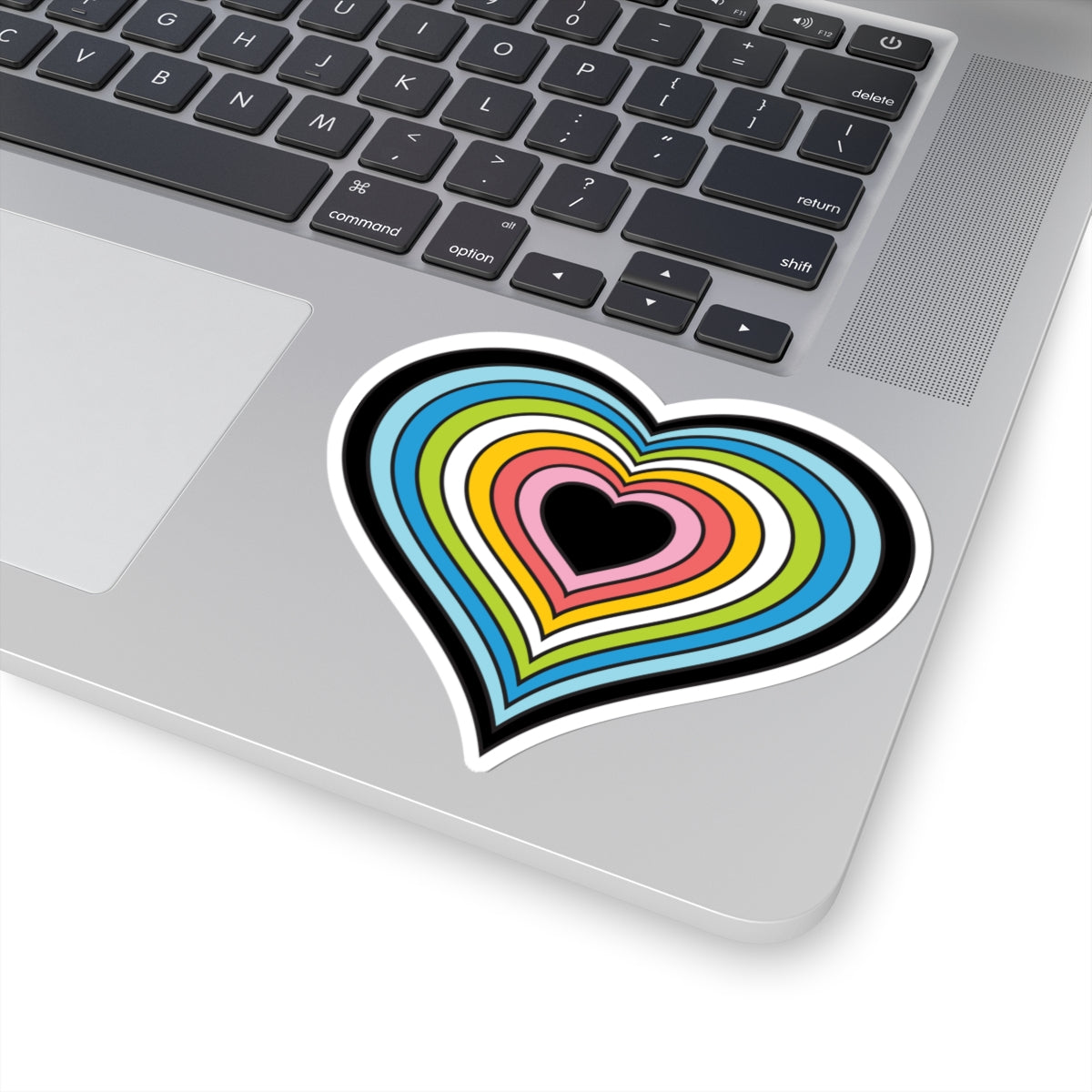 Close-up of the bottom right corner of a silver laptop featuring a 4 inch by 4 inch pride flag heart sticker in a Queer flag style applied to the surface.