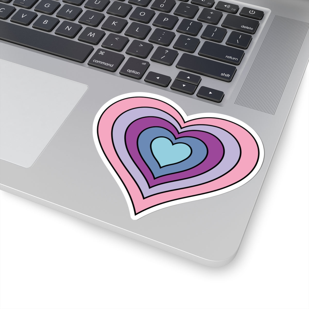 Close-up of the bottom right corner of a silver laptop featuring a 4 inch by 4 inch pride flag heart sticker in an Omnigender flag style applied to the surface.