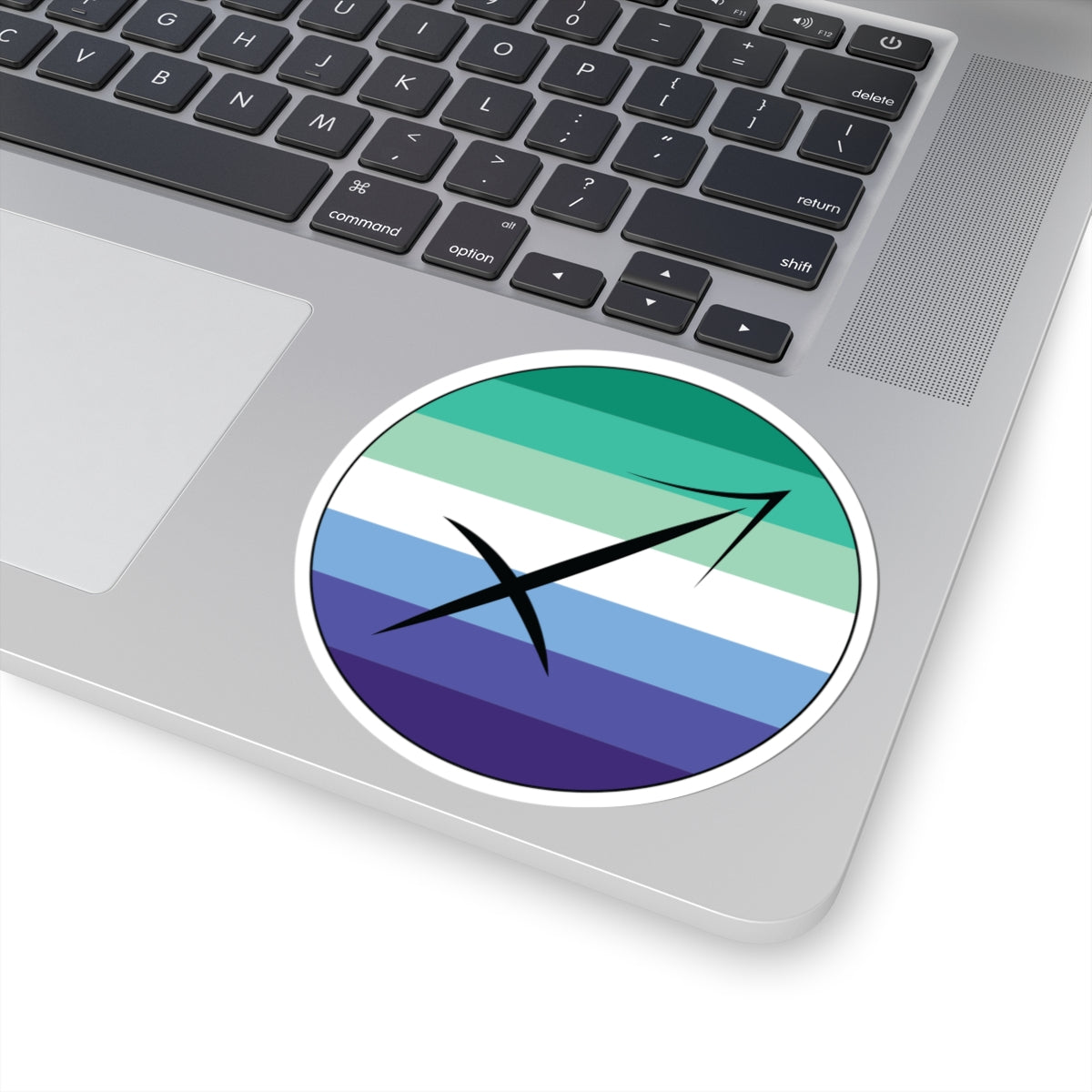 Close-up of the bottom right corner of a silver laptop featuring a 4 inch by 4 inch circular zodiac sticker with a centered Sagittarius glyph on a Gay Men pride flag style background applied to the surface.