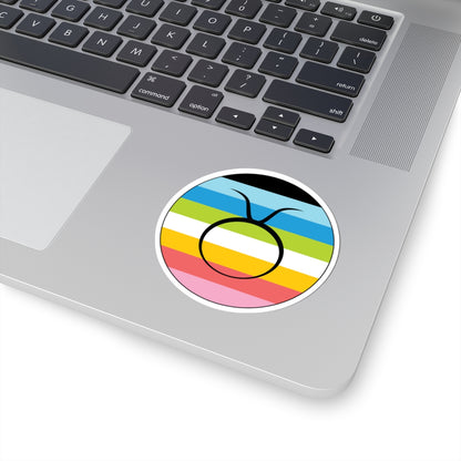 Close-up of the bottom right corner of a silver laptop featuring a 3 inch by 3 inch circular zodiac sticker with a centered Taurus glyph on a Queer pride flag style background applied to the surface.