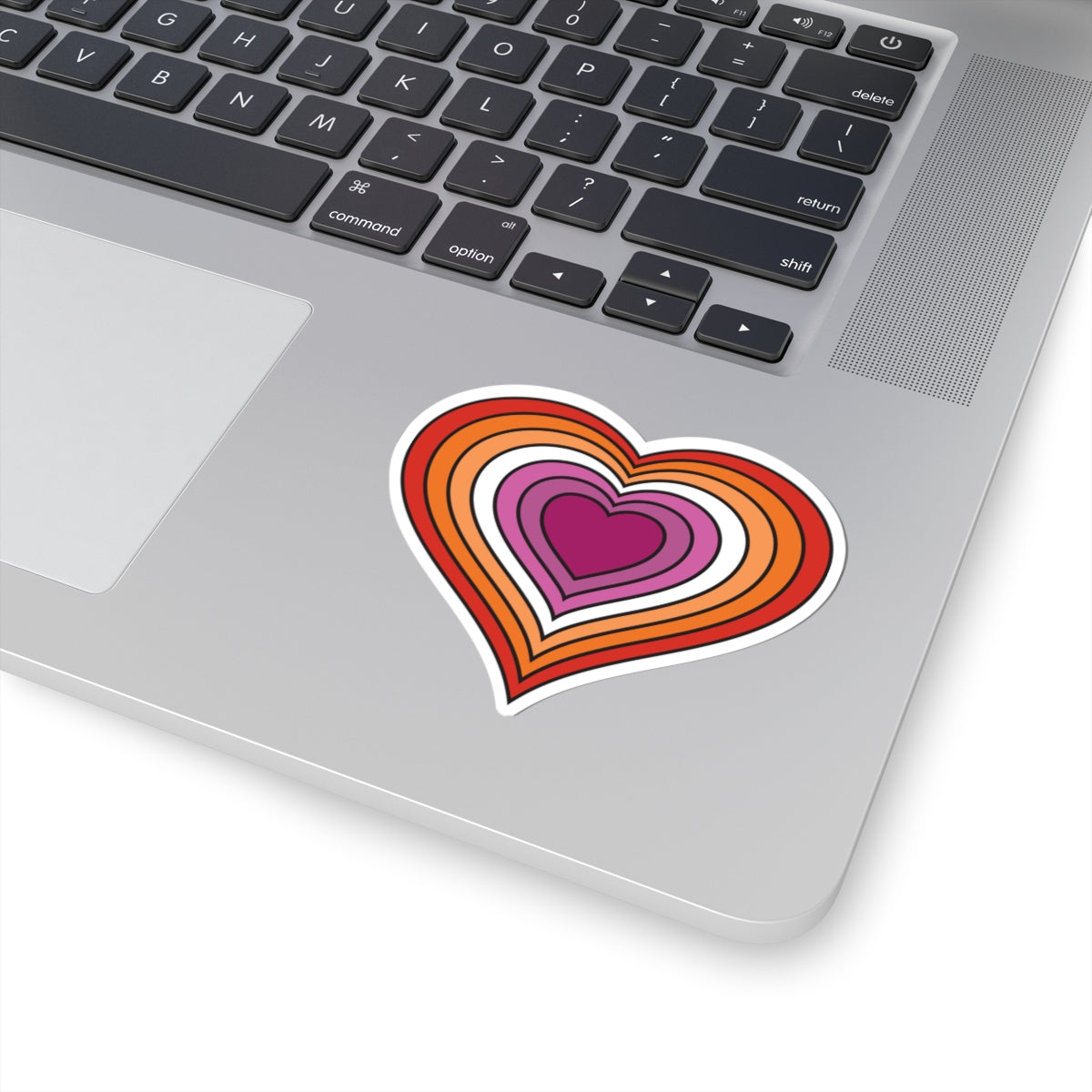 Close-up of the bottom right corner of a silver laptop featuring a 4 inches by 4 inches pride flag heart sticker in a Lesbian pride flag style applied to the surface.