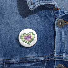 Close-up of a white circular pride button featuring a nested heart design inspired by the Genderfae pride flag in greens, yellow, white, pinks and purple, attached to a blue denim jacket at the right chest with the button clearly visible.