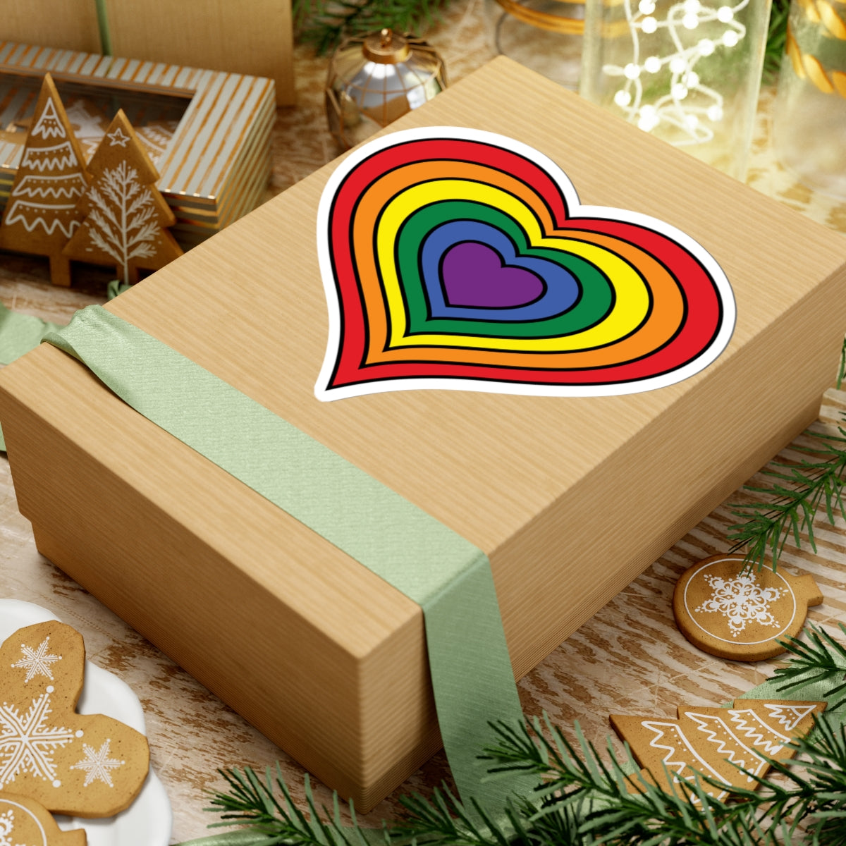 Gift box with a green ribbon on a festive background with cookies and Christmas decorations. Rainbow pride heart sticker centered on the box.