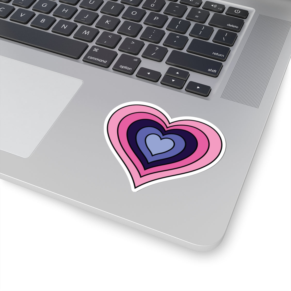 Close-up of the bottom right corner of a silver laptop featuring a 3 inch by 3 inch pride flag heart sticker in an Omnigender flag style applied to the surface.