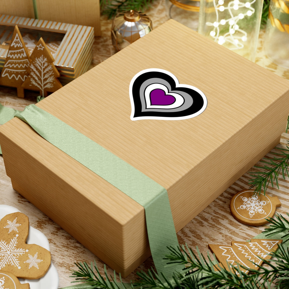 Gift box with a green ribbon on a festive background with cookies and Christmas decorations. Heart-shaped Asexual pride sticker centered on the box.