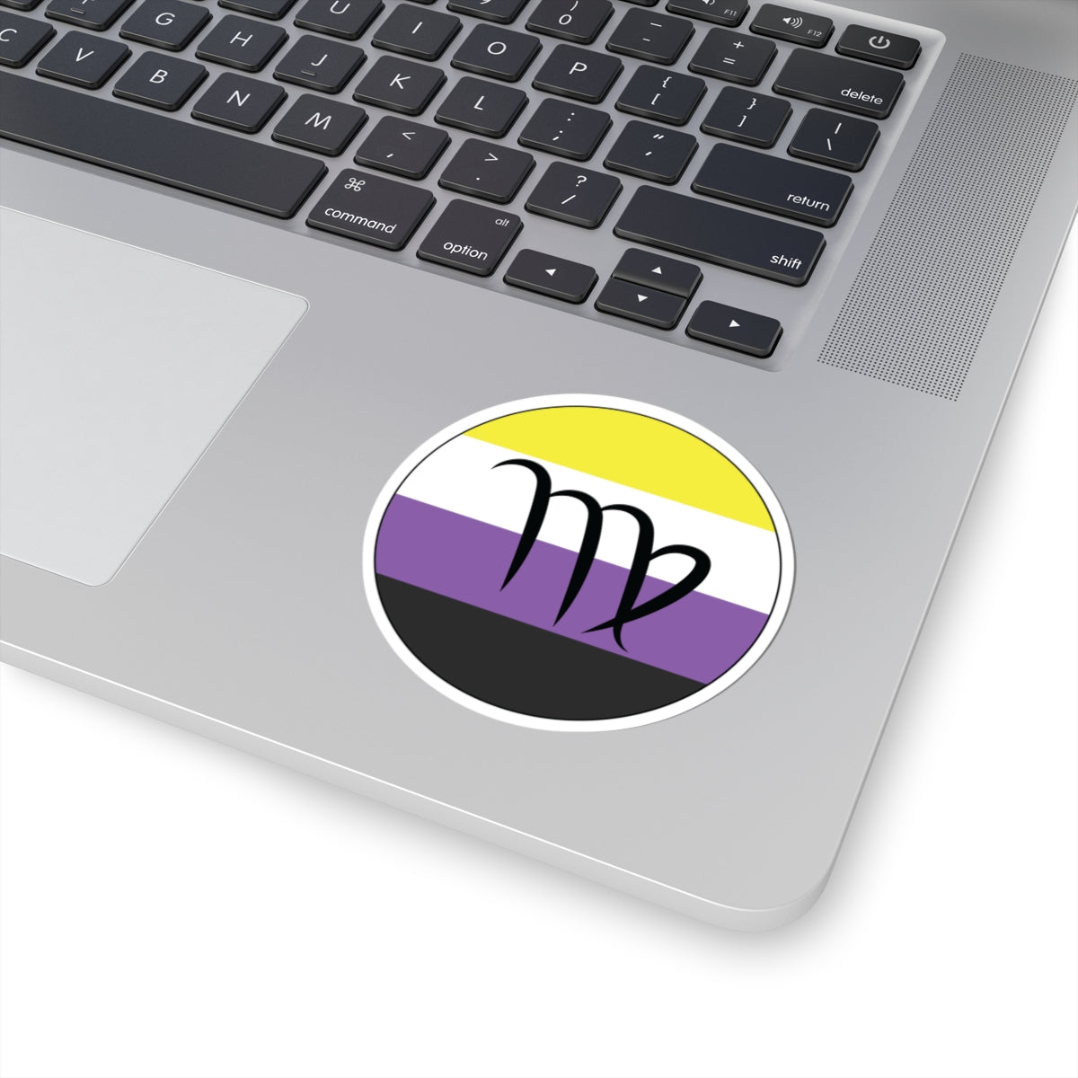 Close-up of the bottom right corner of a silver laptop featuring a 3 inch by 3 inch circular zodiac sticker with a centered Virgo glyph on a Nonbinary pride flag style background applied to the surface.