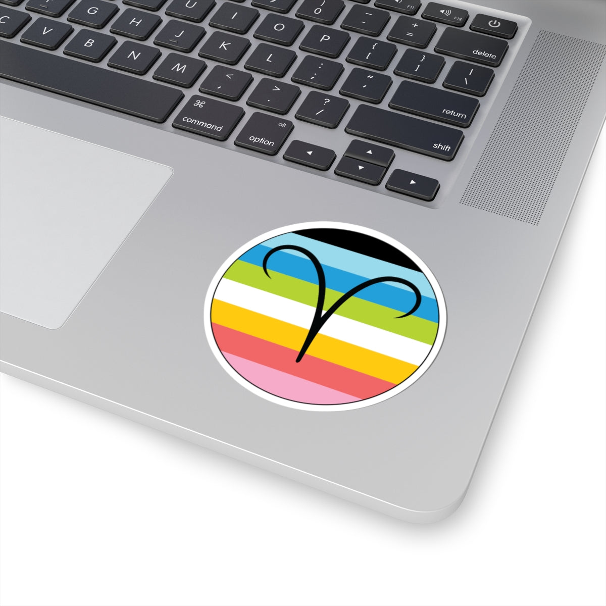 Close-up of the bottom right corner of a silver laptop featuring a 3 inch by 3 inch circular zodiac sticker with a centered Aires glyph on a Queer pride flag style background applied to the surface.