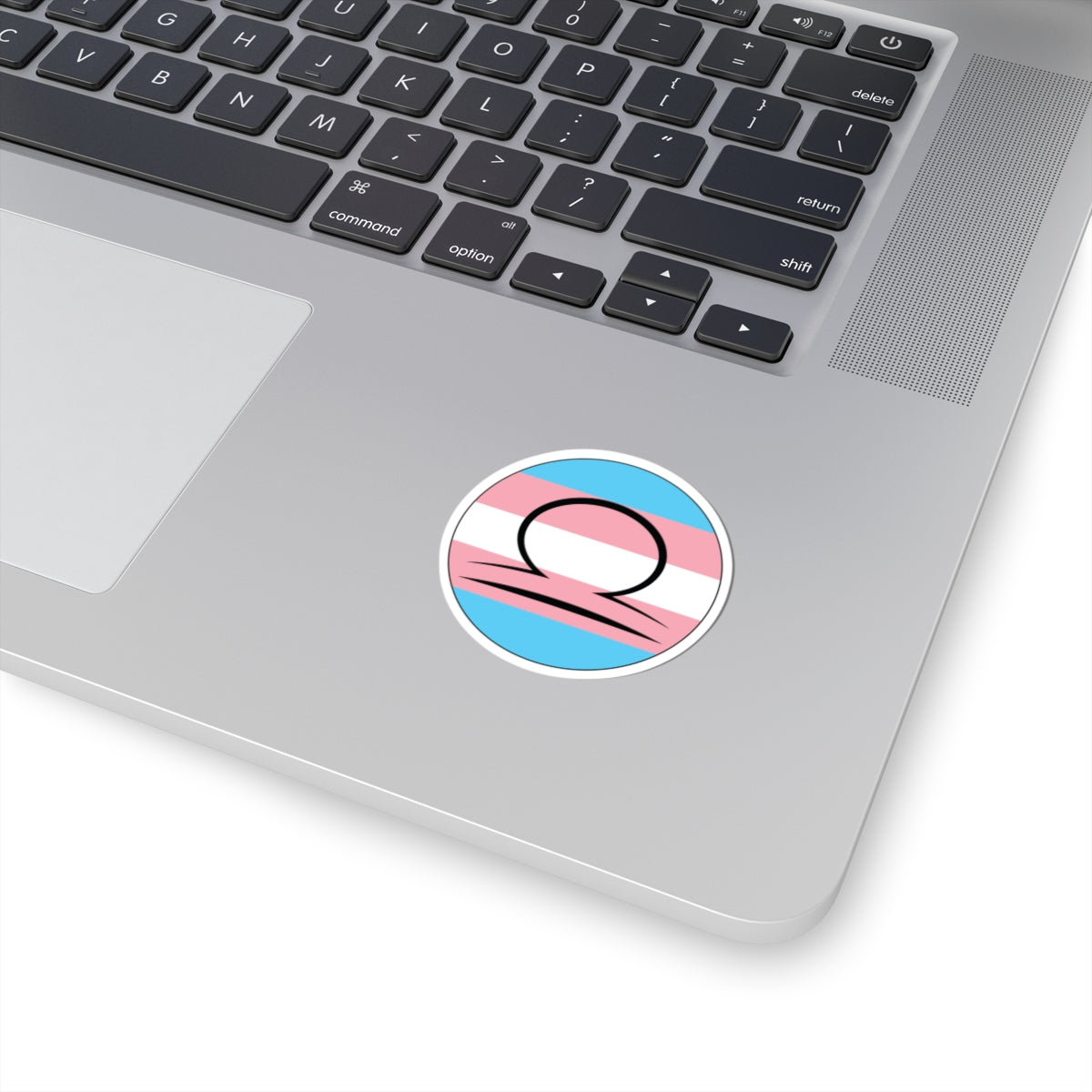Close-up of the bottom right corner of a silver laptop featuring a 2 inch by 2 inch circular zodiac sticker with a centered Libra glyph on a Transgender pride flag style background applied to the surface.