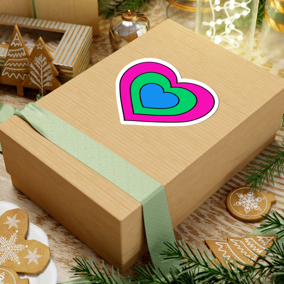 Gift box with a green ribbon on a festive background with cookies and Christmas decorations. Heart-shaped Polysexual pride sticker centered on the box.