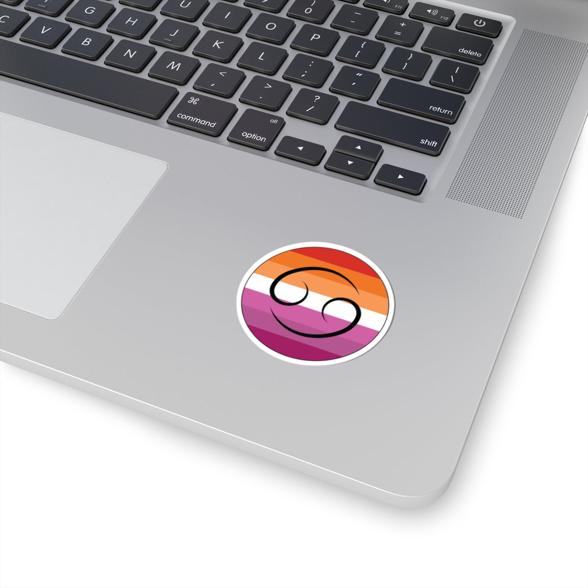 Close-up of the bottom right corner of a silver laptop featuring a 2 inch by 2 inch circular zodiac sticker with a centered Cancer glyph on a Lesbian pride flag style background applied to the surface.