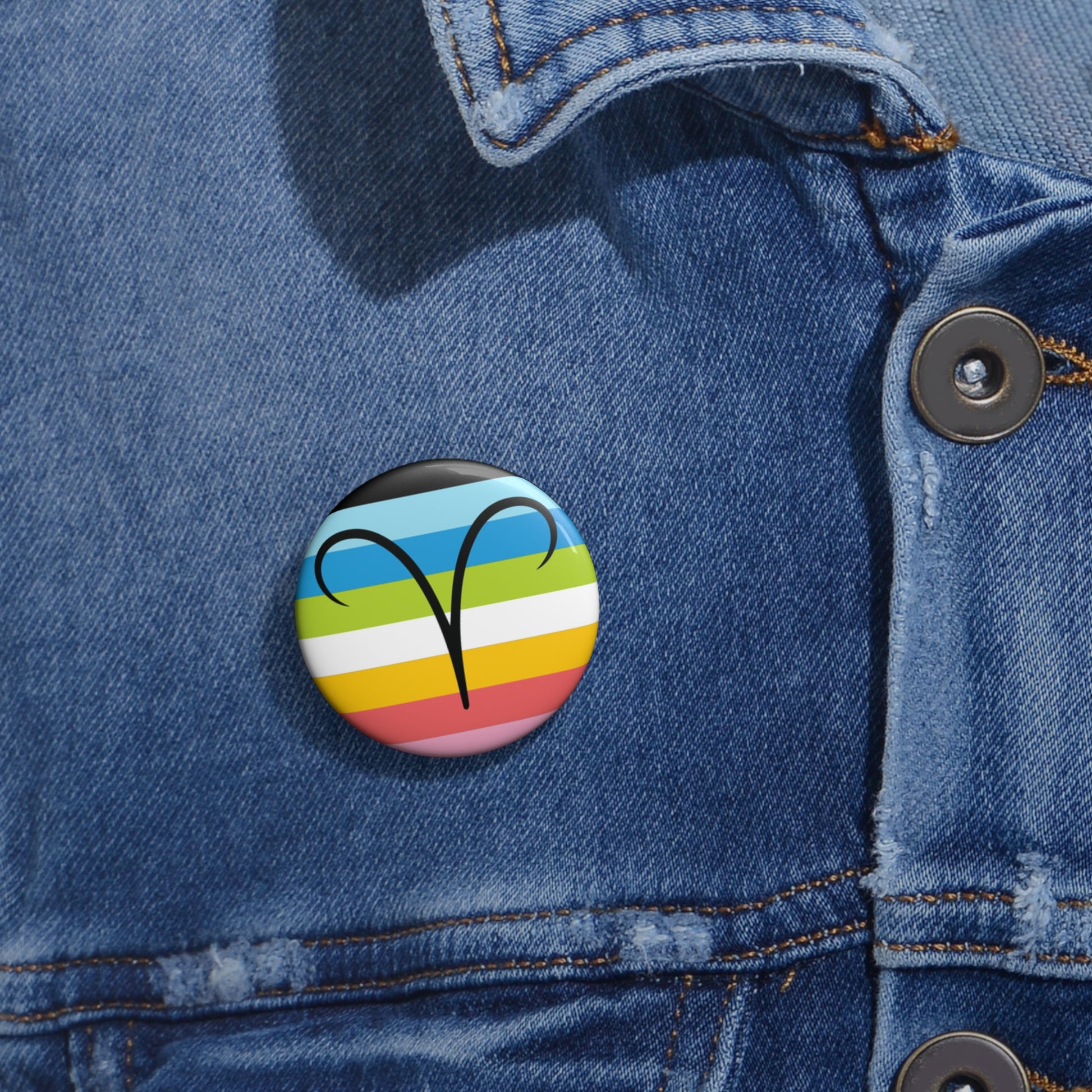 Close-up of a white circular pride button featuring the Aires zodiac glyph centered over a queer pride flag background in pinks, yellow, white, green, blues, and black, attached to a blue denim jacket at the right chest with the button clearly visible.