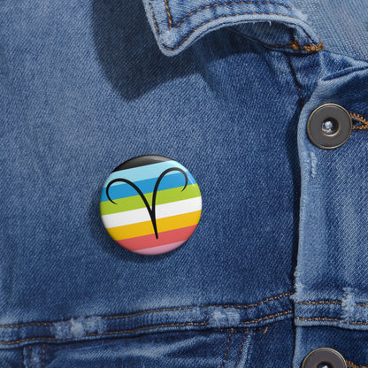 Close-up of a white circular pride button featuring the Aires zodiac glyph centered over a queer pride flag background in pinks, yellow, white, green, blues, and black, attached to a blue denim jacket at the right chest with the button clearly visible.
