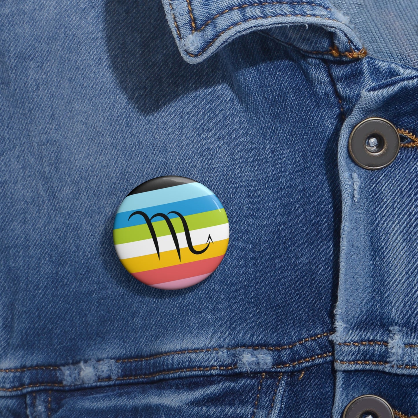 Close-up of a white circular pride button featuring the Scorpio zodiac glyph centered over a queer pride flag background in pinks, yellow, white, green, blues, and black, attached to a blue denim jacket at the right chest with the button clearly visible.