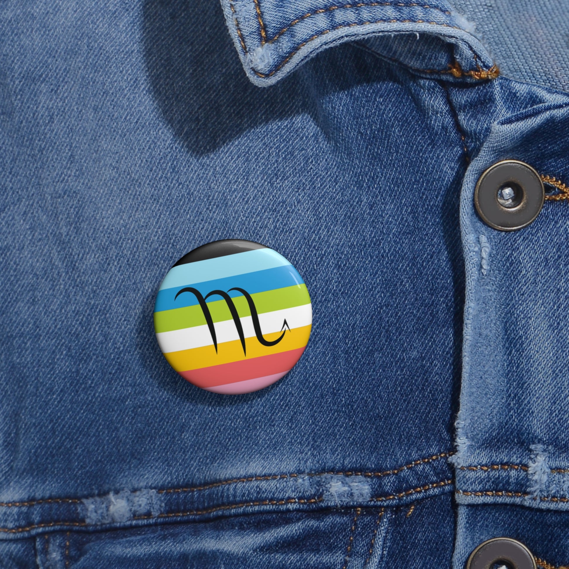 Close-up of a white circular pride button featuring the Scorpio zodiac glyph centered over a queer pride flag background in pinks, yellow, white, green, blues, and black, attached to a blue denim jacket at the right chest with the button clearly visible.
