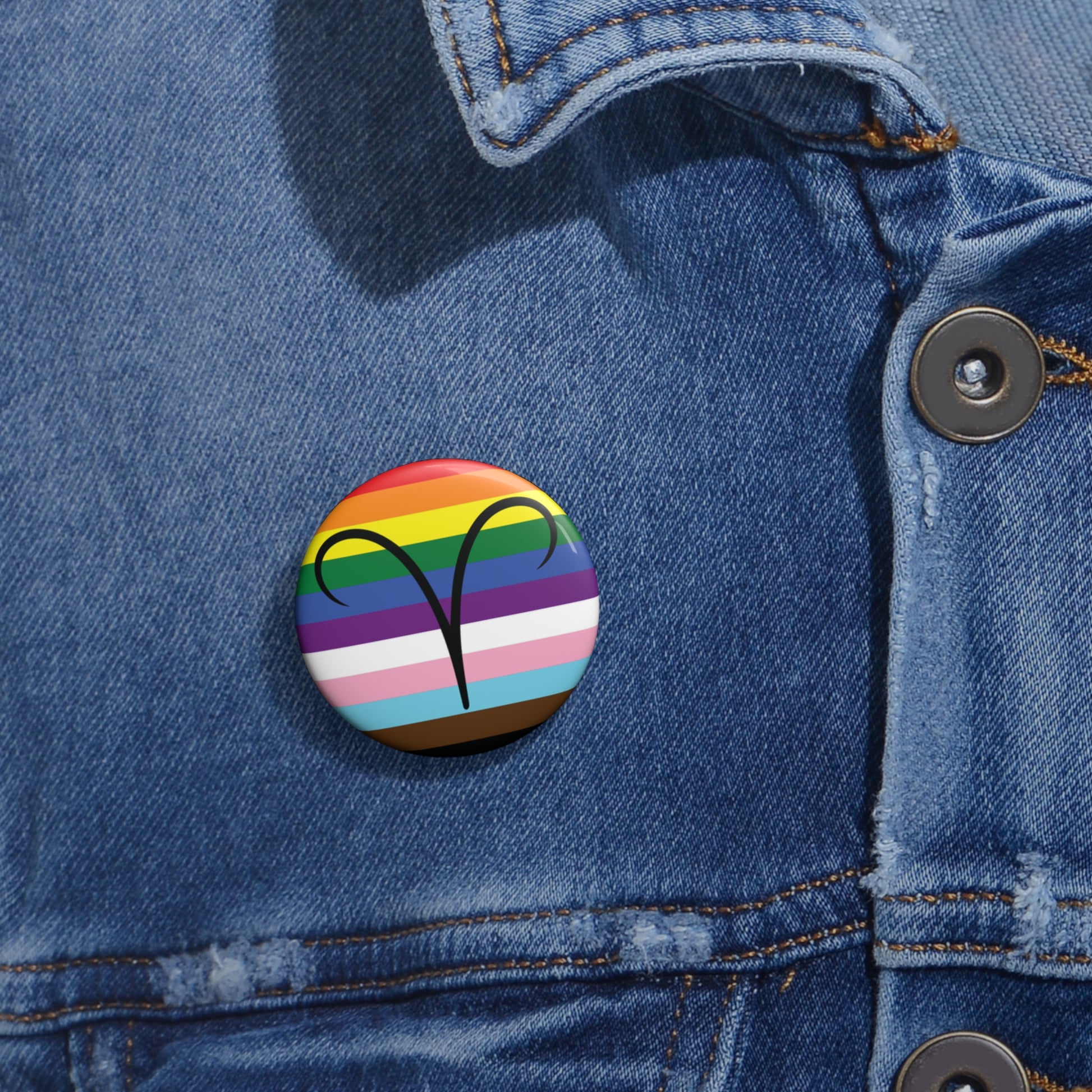 Close-up of a white circular pride button featuring the Aires zodiac glyph centered over a Progress Pride flag background in rainbow stripes with black, brown, light blue, pink, and white elements, attached to a blue denim jacket at the right chest with the button clearly visible.