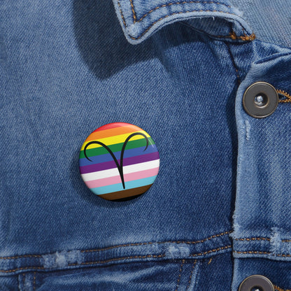 Close-up of a white circular pride button featuring the Aires zodiac glyph centered over a Progress Pride flag background in rainbow stripes with black, brown, light blue, pink, and white elements, attached to a blue denim jacket at the right chest with the button clearly visible.