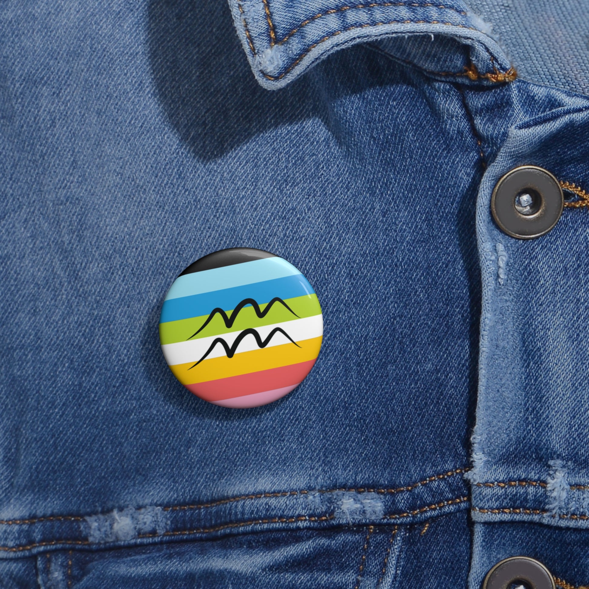 Close-up of a white circular pride button featuring the Aquarius zodiac glyph centered over a queer pride flag background in pinks, yellow, white, green, blues, and black, attached to a blue denim jacket at the right chest with the button clearly visible.