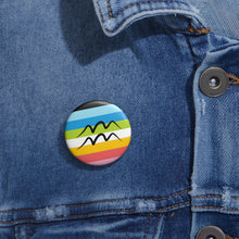 Close-up of a white circular pride button featuring the Aquarius zodiac glyph centered over a queer pride flag background in pinks, yellow, white, green, blues, and black, attached to a blue denim jacket at the right chest with the button clearly visible.