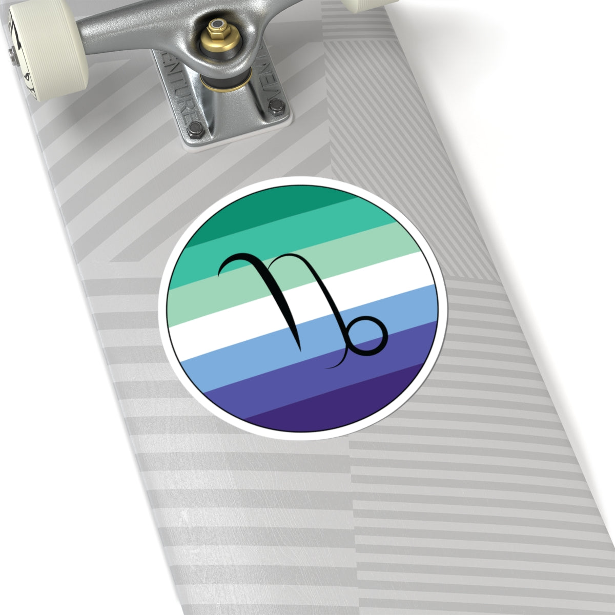 Close-up of the bottom of a silver skateboard featuring a 6 inch by 6 inch circular zodiac sticker with a centered Capricorn glyph on a Gay Men pride flag style background applied to the surface.
