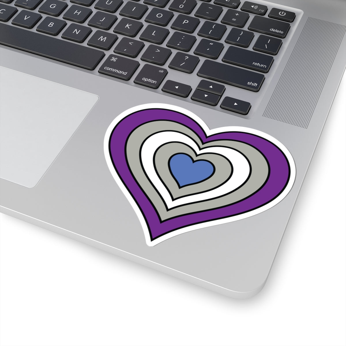 Close-up of the bottom right corner of a silver laptop featuring a 4 inch by 4 inch pride flag heart sticker in a Greysexual flag style applied to the surface.