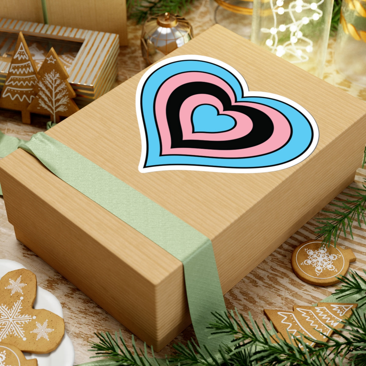 Gift box with a green ribbon on a festive background with cookies and Christmas decorations. Heart-shaped Transfeminine pride sticker centered on the box.