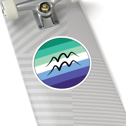 Close-up of the bottom of a silver skateboard featuring a 6 inch by 6 inch circular zodiac sticker with a centered Aquarius glyph on a Gay Men pride flag style background applied to the surface.
