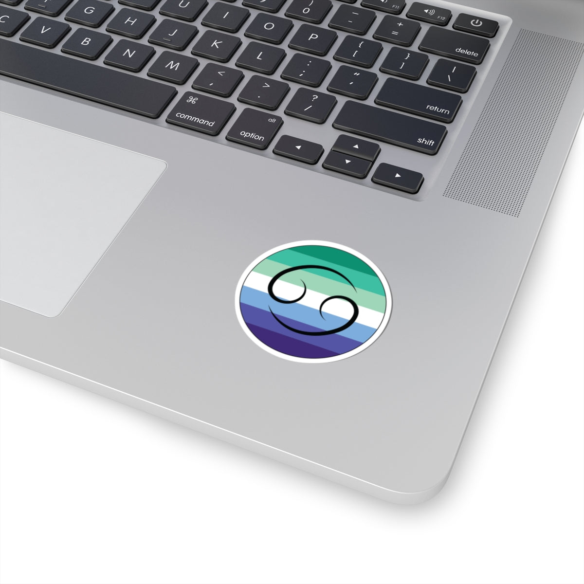 Close-up of the bottom right corner of a silver laptop featuring a 2 inch by 2 inch circular zodiac sticker with a centered Cancer glyph on a Gay Men pride flag style background applied to the surface.