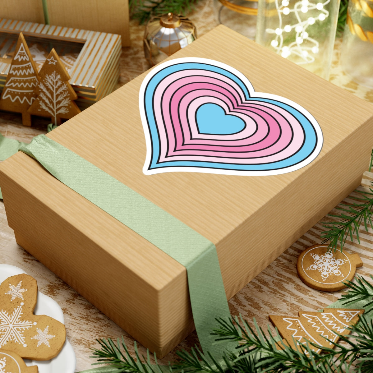 Gift box with a green ribbon on a festive background with cookies and Christmas decorations. Heart-shaped Transfeminine pride sticker centered on the box.