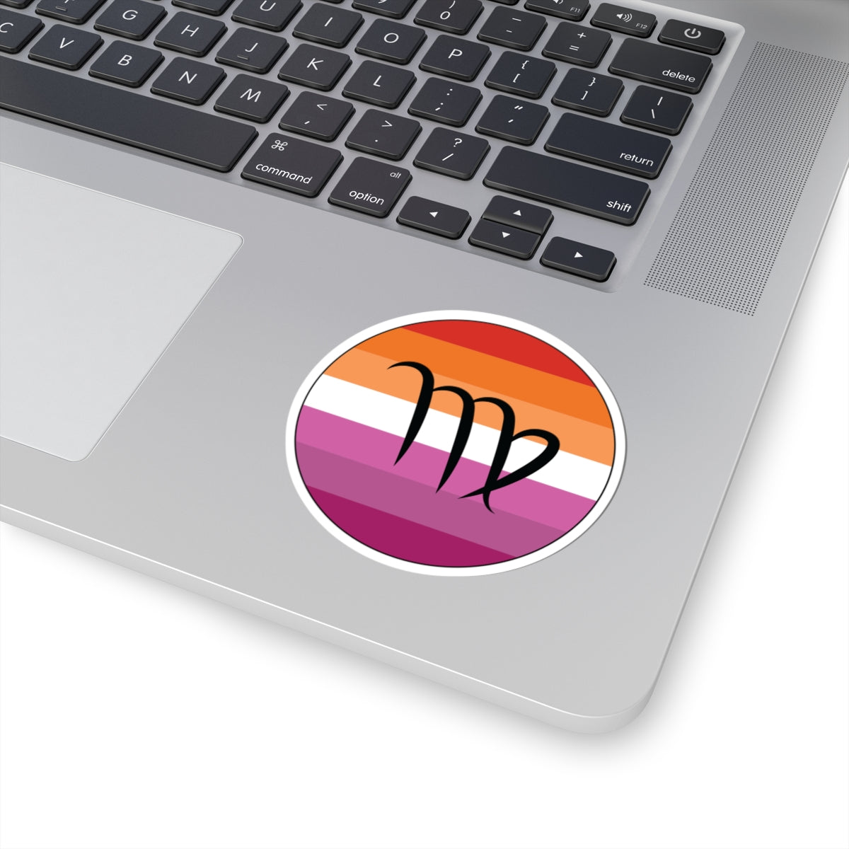 Close-up of the bottom right corner of a silver laptop featuring a 3 inch by 3 inch circular zodiac sticker with a centered Virgo glyph on a Lesbian pride flag style background applied to the surface.