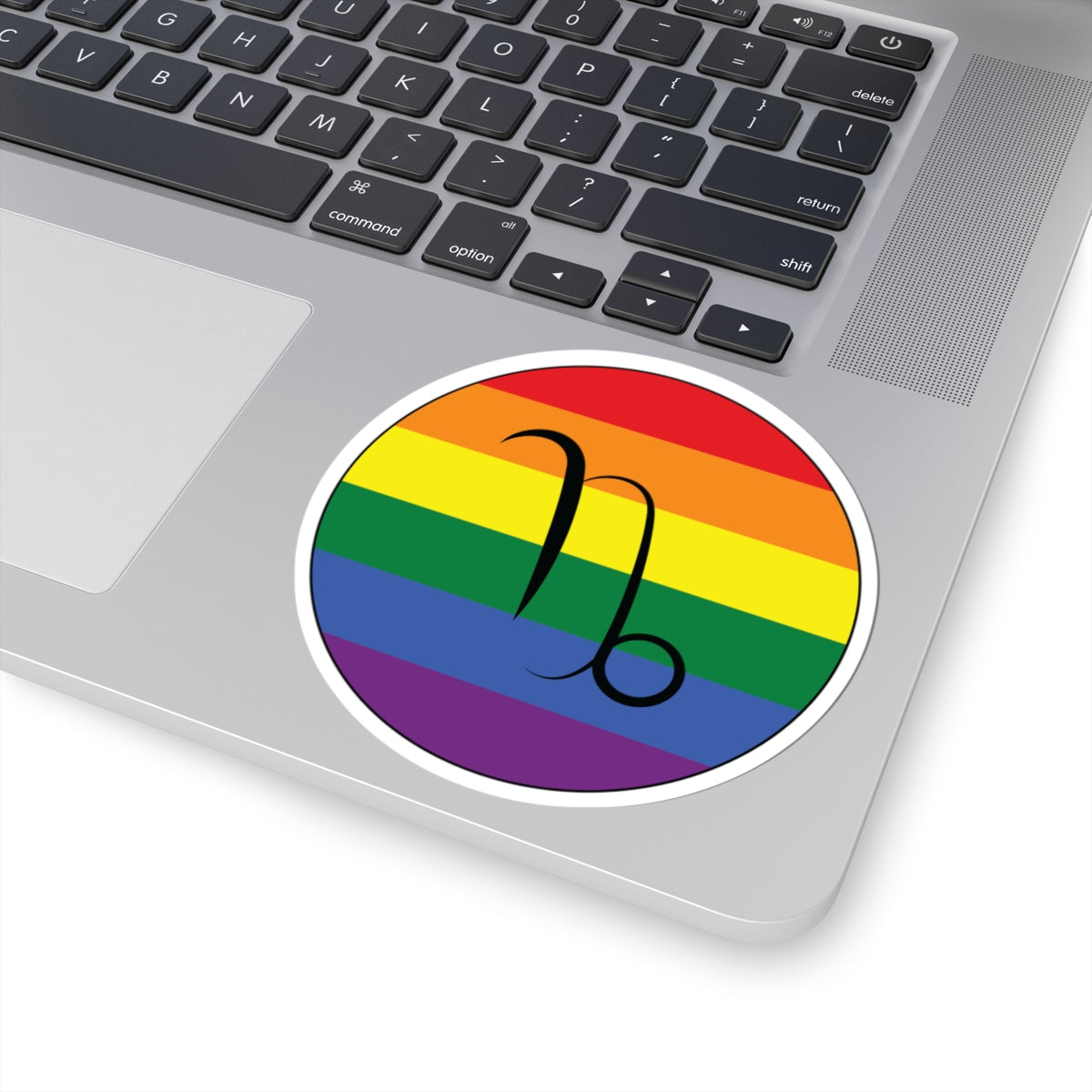 Close-up of the bottom right corner of a silver laptop featuring a 4 inch by 4 inch circular zodiac sticker with a centered Capricorn glyph on a Rainbow pride flag style background applied to the surface.