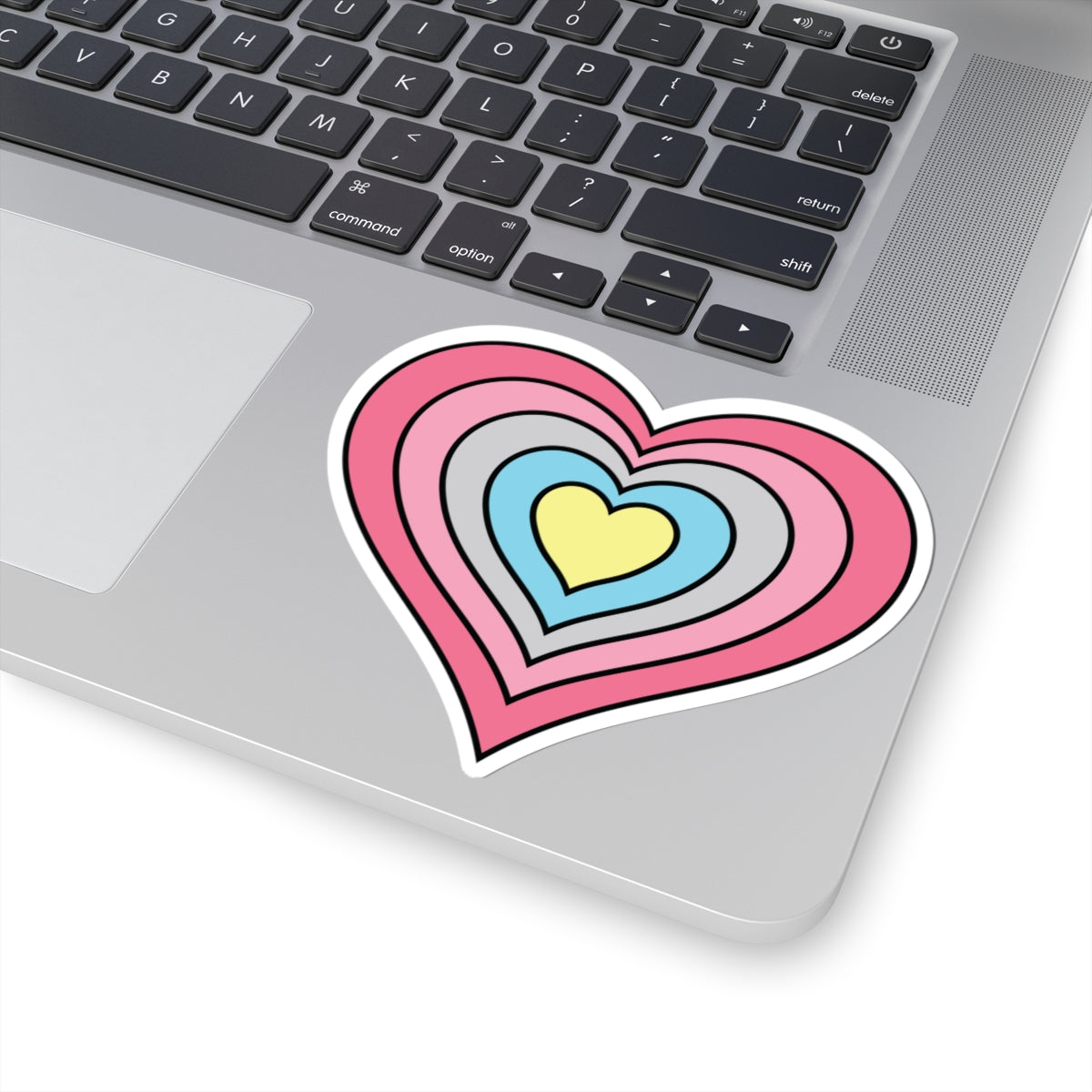 Close-up of the bottom right corner of a silver laptop featuring a 4 inch by 4 inch pride flag heart sticker in a Genderflux flag style applied to the surface.