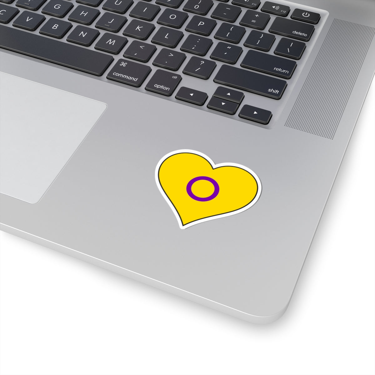 Close-up of the bottom right corner of a silver laptop featuring a 2 inch by 2 inch pride flag heart sticker in an Intersex flag style applied to the surface.