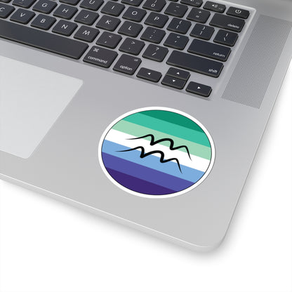 Close-up of the bottom right corner of a silver laptop featuring a 3 inch by 3 inch circular zodiac sticker with a centered Aquarius glyph on a Gay Men pride flag style background applied to the surface.