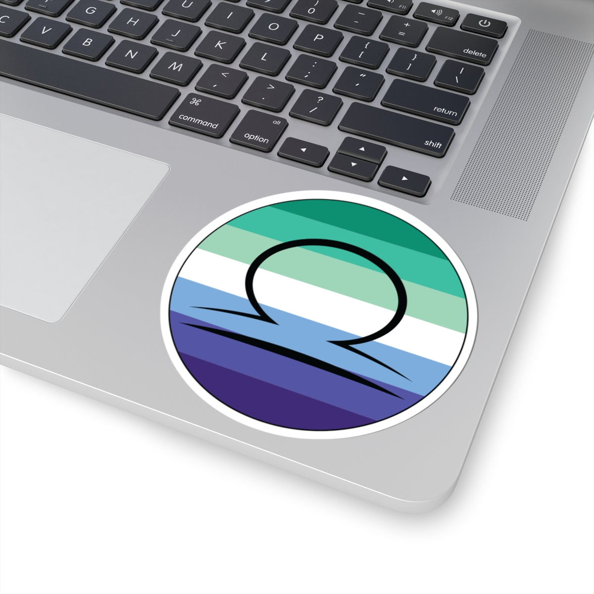 Close-up of the bottom right corner of a silver laptop featuring a 4 inch by 4 inch circular zodiac sticker with a centered Libra glyph on a Gay Men pride flag style background applied to the surface.