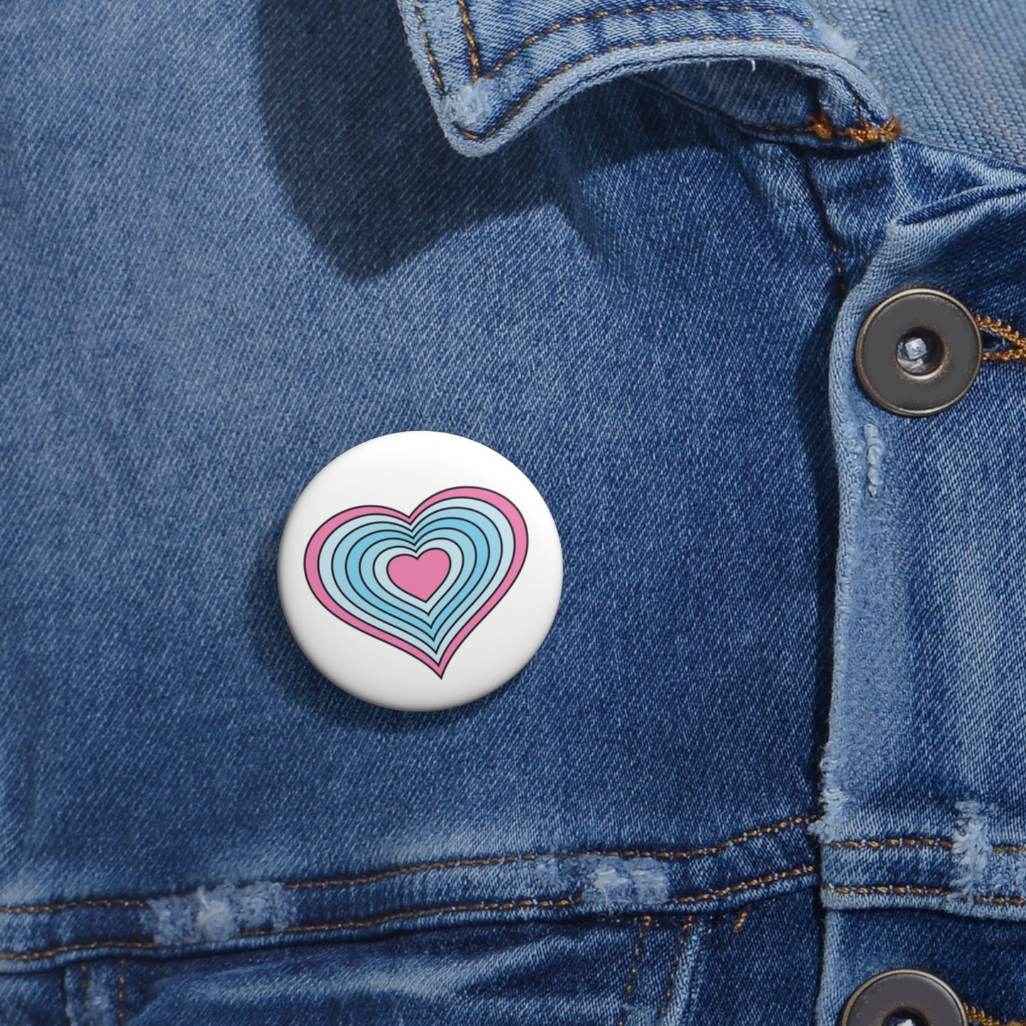 Close-up of a white circular pride button featuring a nested heart design inspired by the Transmasc pride flag in pink and blues, attached to a blue denim jacket at the right chest with the button clearly visible.