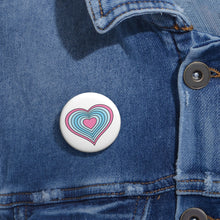 Close-up of a white circular pride button featuring a nested heart design inspired by the Transmasc pride flag in pink and blues, attached to a blue denim jacket at the right chest with the button clearly visible.