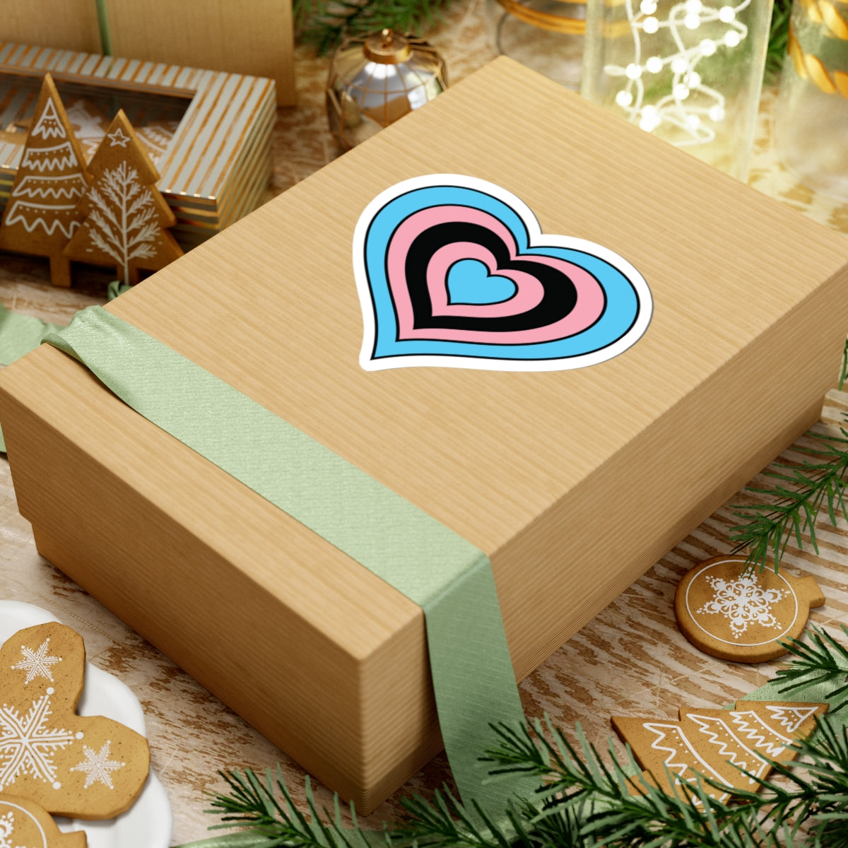 Gift box with a green ribbon on a festive background with cookies and Christmas decorations. Heart-shaped Transfeminine pride sticker centered on the box.