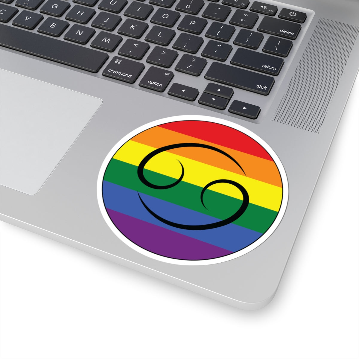 Close-up of the bottom right corner of a silver laptop featuring a 4 inch by 4 inch circular zodiac sticker with a centered Cancer glyph on a Rainbow pride flag style background applied to the surface.