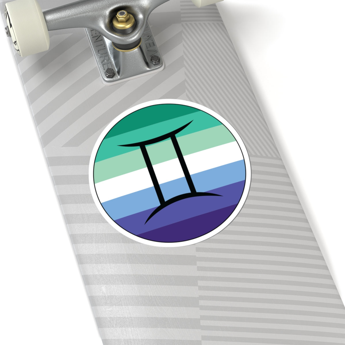 Close-up of the bottom of a silver skateboard featuring a 6 inch by 6 inch circular zodiac sticker with a centered Gemini glyph on a Gay Men pride flag style background applied to the surface.
